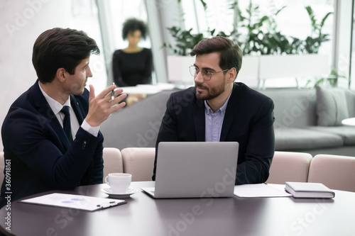 Young business man explaining to senior colleague new strategy. Serious advisor consulting client using computer, office manager listen to coworkers plan. Insurer or salesman giving advice to client