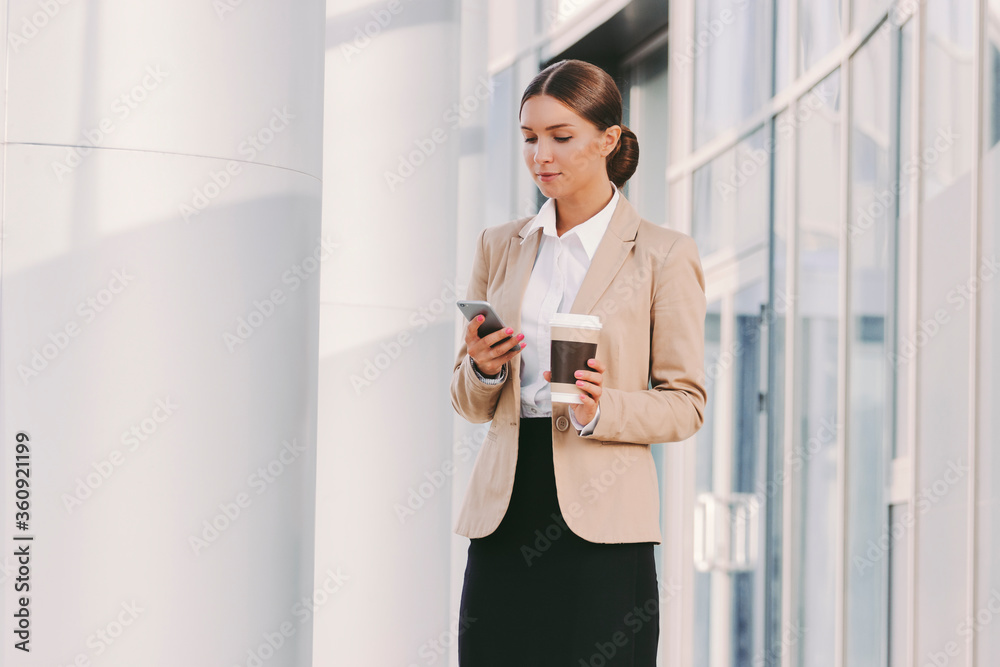 Portrait young beautiful business woman in stylish suit use smart phone and drink coffee outdoors. Attractive girl professional entrepreneur hold paper coffee cup and mobile phone in hand. Lunch break