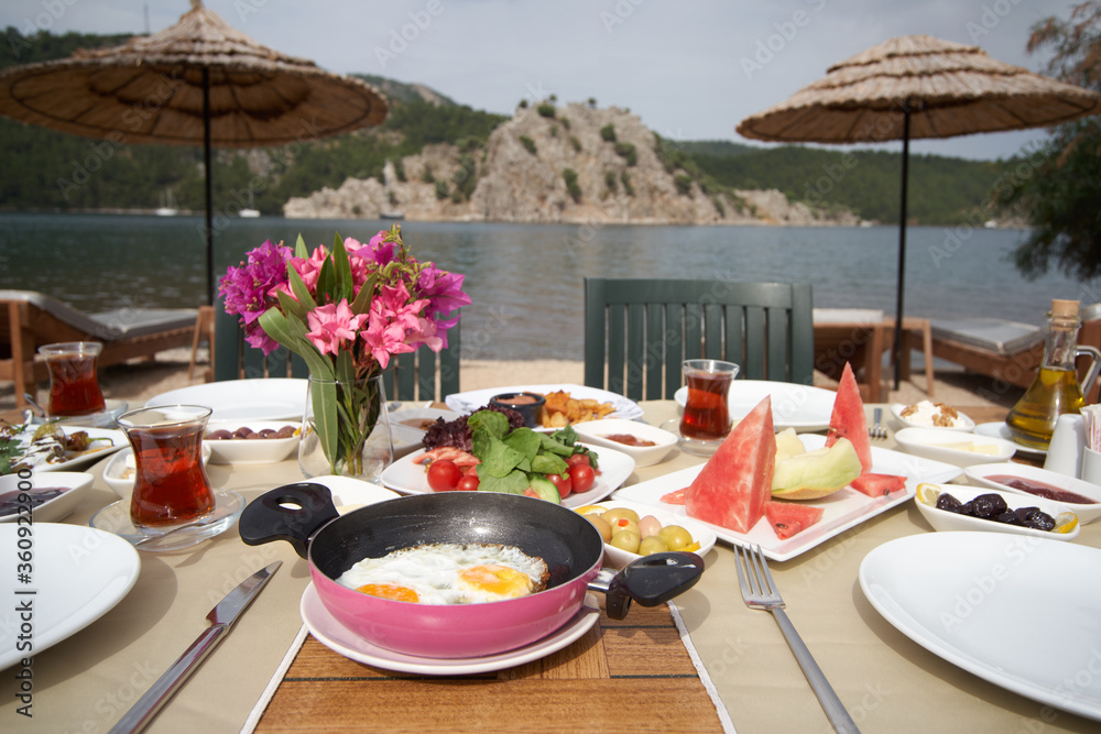 A breakfast table with fried egg, tomato and fruits placed in front of sea view