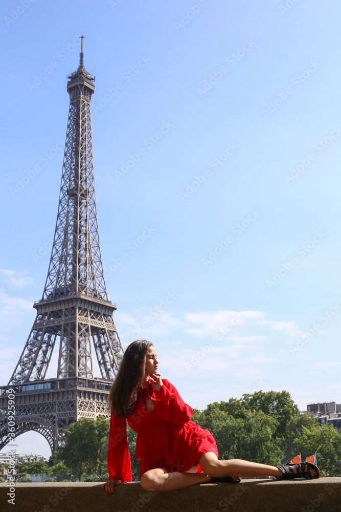 Young girl in red dress sits alone in front of Eiffel Tower in Paris on summer sunny day