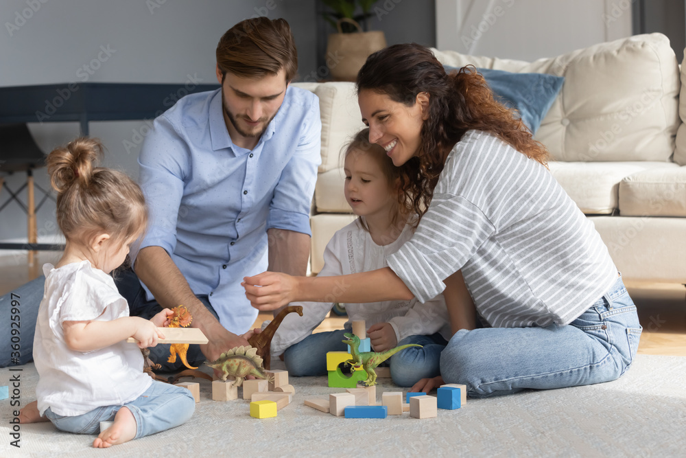 Happy parents with two little daughters playing with toys colorful wooden blocks and animals, sitting on warm floor in living room, happy young family with cute preschool kids enjoying weekend Happy parents with two little daughters playing with toys colorful wooden blocks and animals, sitting on warm floor in living room, happy young family with cute preschool kids enjoying weekend