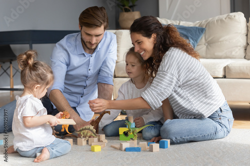 Happy parents with two little daughters playing with toys colorful wooden blocks and animals, sitting on warm floor in living room, happy young family with cute preschool kids enjoying weekend