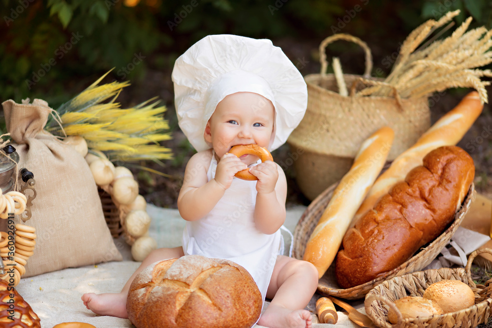 Healthy eating concept. Happy boy is cooking on nature on a sunny summer day. Toddler baker on a picnic eats bread and bagels in a white apron and hat Healthy eating concept. Happy boy is cooking on nature on a sunny summer day. Toddler baker on a picnic eats bread and bagels in a white apron and hat