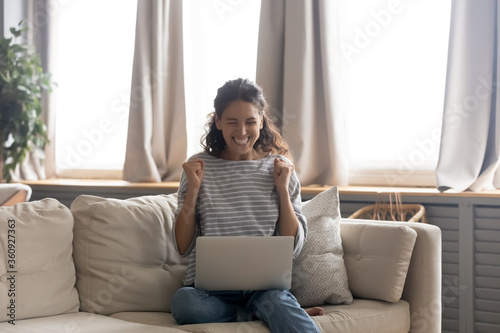 Excited young woman looking at laptop screen, celebrating win, showing yes gesture, sitting on cozy couch in living room at home, rejoicing success, reading good news in email, online lottery victory photo