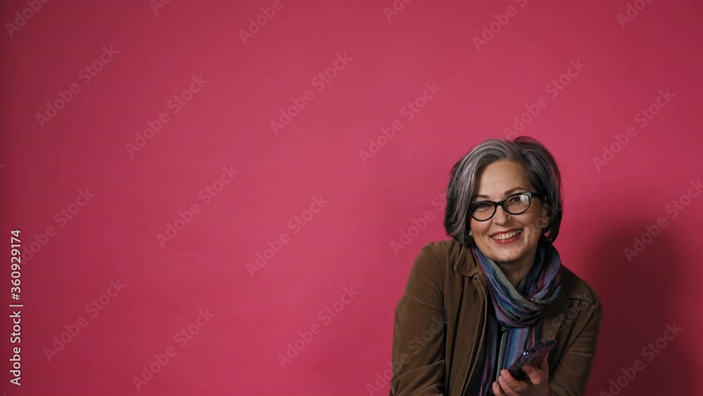 Graying pretty woman take phone down smiles. Smart mature lady in eyeglasses and brown jacket on pink background. Text space at left side. Prores 422. Graying pretty woman take phone down smiles. Smart mature lady in eyeglasses and brown jacket on pink background. Text space at left side. Prores 422.
