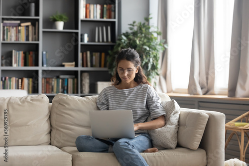 Focused beautiful woman wearing glasses using laptop in living room, sitting on cozy couch, interested girl looking at computer screen, browsing apps, chatting or studying, working online