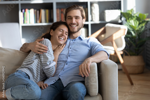 Happy young couple hugging, laughing at joke, sitting on cozy couch in living room, happy overjoyed woman and man cuddling, having fun together, enjoying leisure time, lazy weekend at home