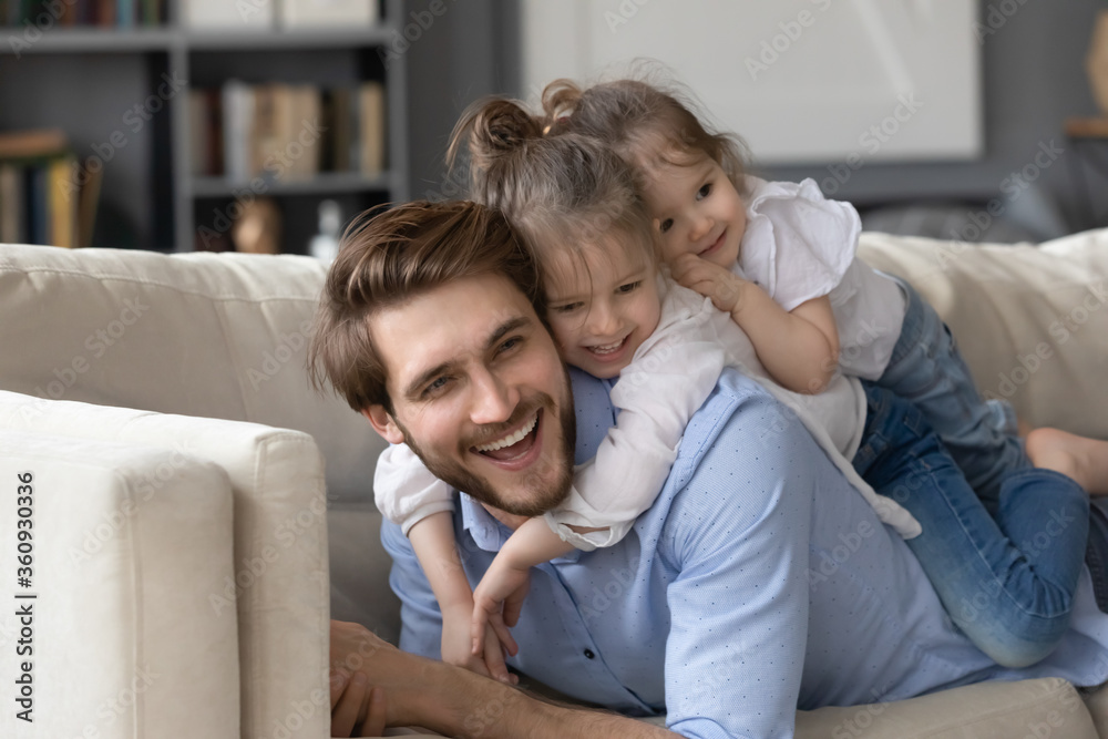Two cute little girls lying on smiling father back, relaxing on cozy couch together, enjoying leisure time, lazy weekend, happy young dad and affectionate preschool daughters having fun at home Two cute little girls lying on smiling father back, relaxing on cozy couch together, enjoying leisure time, lazy weekend, happy young dad and affectionate preschool daughters having fun at home