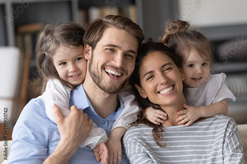 Head shot portrait two little daughters piggy backing happy parents, posing for funny family photo, smiling beautiful mother and father hugging with adorable kids at home, enjoying leisure time