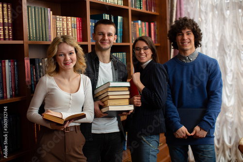 Four smiling students with stacks of books in their hands rejoice that they will have the last exams at the university against  the background of the bookcase in the library
