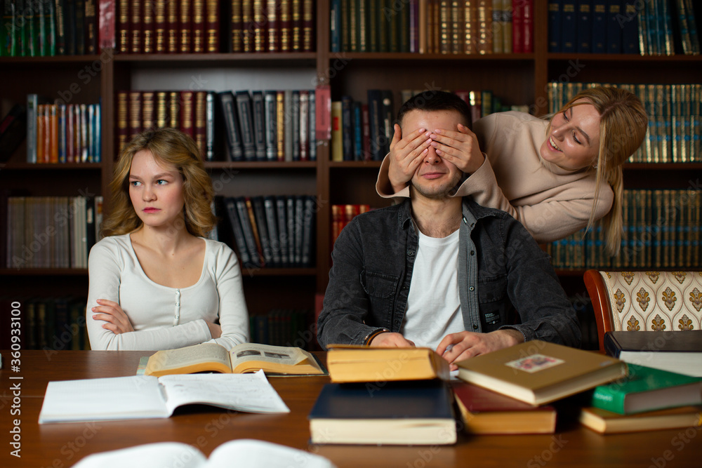 A girl in a beige blouse smiling closed the eyes of a guy in a gray shirt with her hands, and a girl in a white blouse on the side offended looks to the side against the background of bookcases A girl in a beige blouse smiling closed the eyes of a guy in a gray shirt with her hands, and a girl in a white blouse on the side offended looks to the side against the background of bookcases