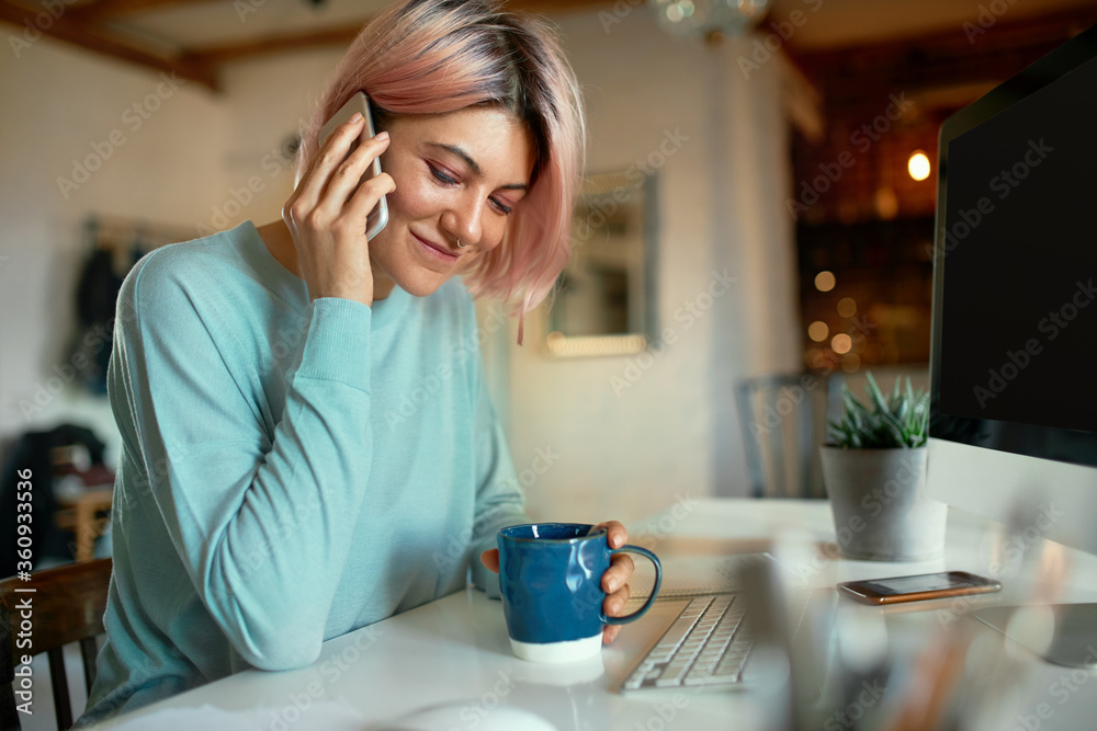 Fashionable young female copywriter with pink hair and facial piercing sitting at her workplace in front of desktop computer, holding cup, drinking coffee and having phone conversation, smiling Fashionable young female copywriter with pink hair and facial piercing sitting at her workplace in front of desktop computer, holding cup, drinking coffee and having phone conversation, smiling