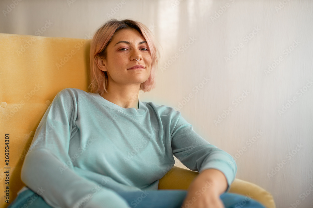 Horizontal shot of stylish pretty girl with bob pink hairstyle and nose ring sitting comfortably in armchair, looking at camera with carefree relaxed smile. People, lifestyle, leisure and relaxation Horizontal shot of stylish pretty girl with bob pink hairstyle and nose ring sitting comfortably in armchair, looking at camera with carefree relaxed smile. People, lifestyle, leisure and relaxation