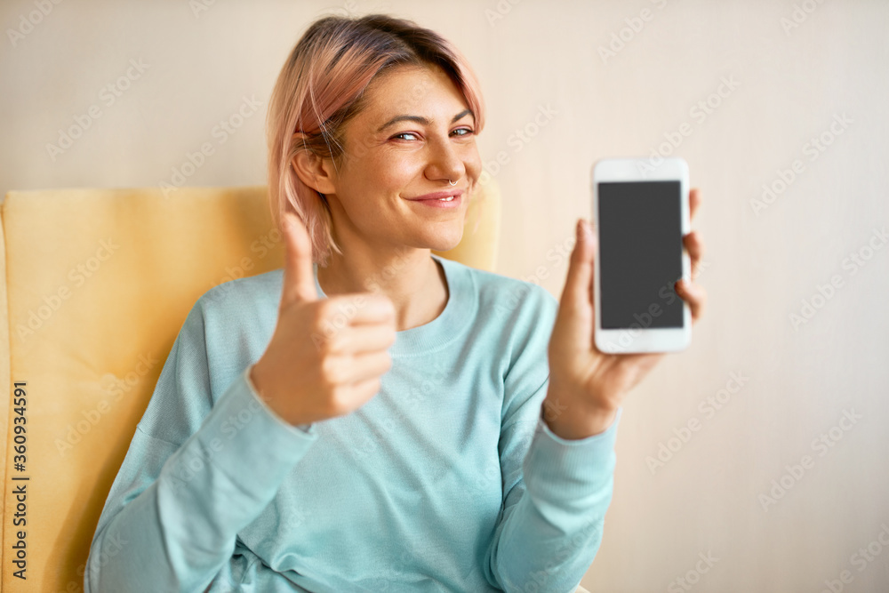 Charming happy young woman with pinkish hair holding smart phone with blank black display with copy space for your advertising content, making thumbs up gesture as sign of appoval, winking at camera