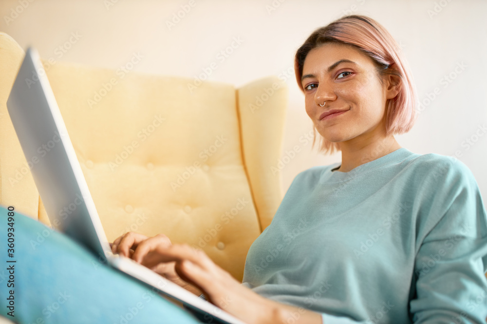 Low angle view of charming stylish student girl doing homework using generic laptop, sitting on sofa, keyboarding, using high speed wireless internet connection. Technology and electronic gadgets Low angle view of charming stylish student girl doing homework using generic laptop, sitting on sofa, keyboarding, using high speed wireless internet connection. Technology and electronic gadgets
