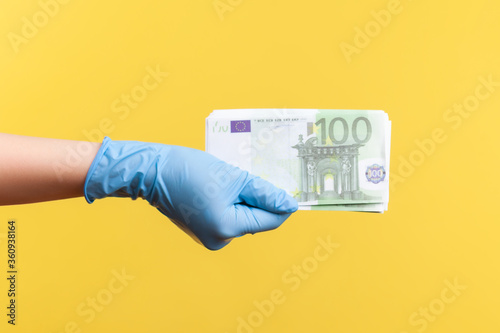 Profile side view closeup of human hand in blue surgical gloves holding and showing fan of European union Euro money in hand. indoor, studio shot, isolated on yellow background.