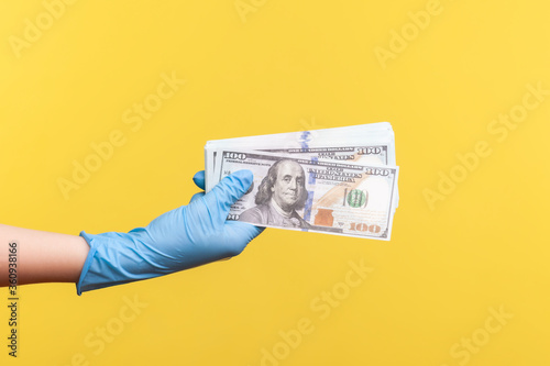 Profile side view closeup of human hand in blue surgical gloves holding and showing fan of american dollar money in hand. indoor, studio shot, isolated on yellow background.
