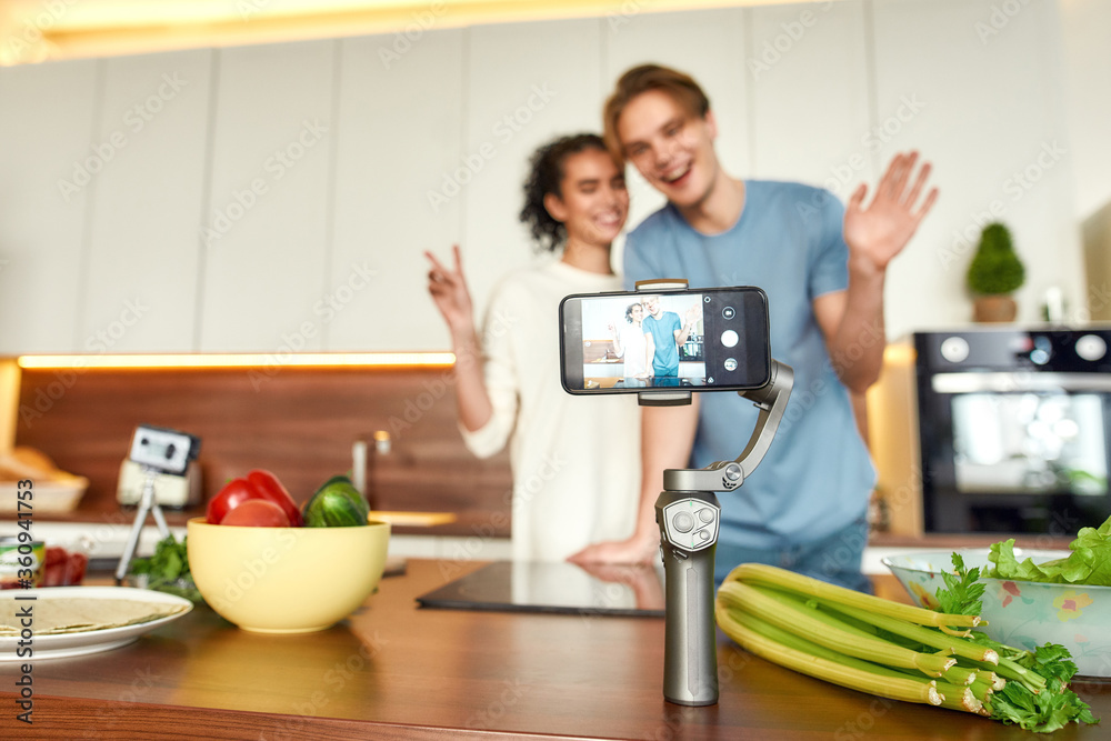 Happy couple, vegetarians smiling, recording a video on smartphone while going to prepare a healthy meal in the kitchen together. Vegetarianism, healthy food, blog concept Happy couple, vegetarians smiling, recording a video on smartphone while going to prepare a healthy meal in the kitchen together. Vegetarianism, healthy food, blog concept