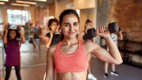 Young female trainer looking at camera while working out using dumbbell in gym together with kids, teenagers. Sport, healthy lifestyle, physical education concept