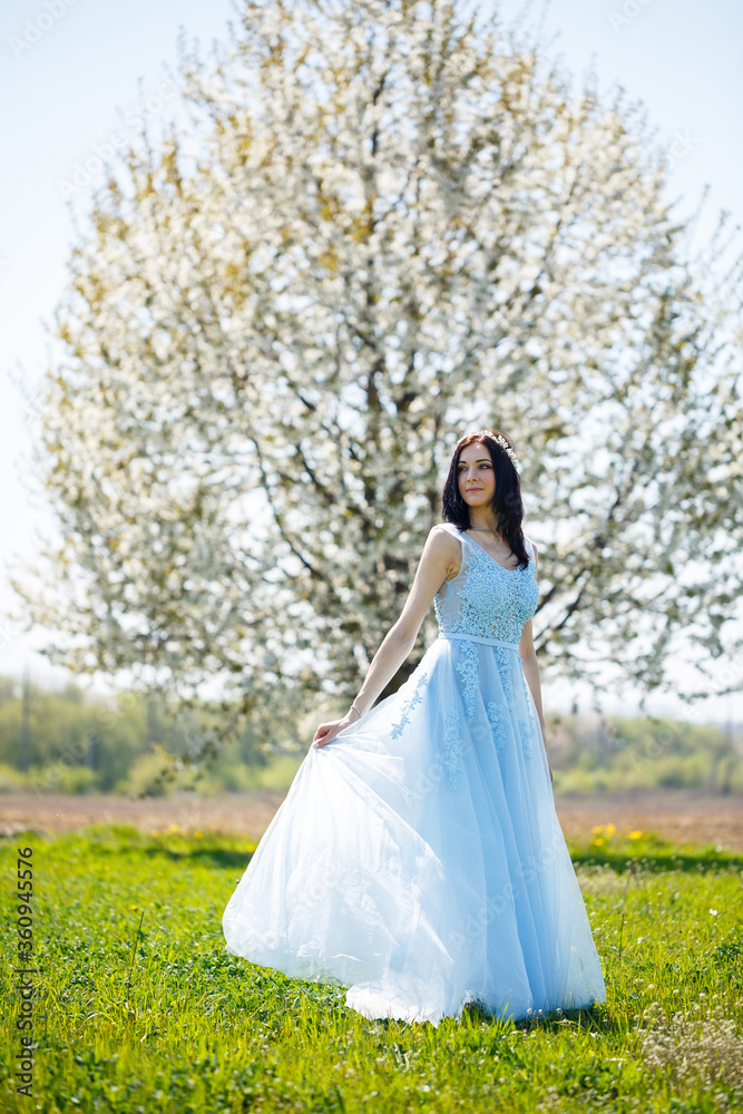 Portrait of a girl in a light summer blue long dress adorned in her hair against a blossoming tree. Portrait of a girl in a light summer blue long dress adorned in her hair against a blossoming tree.