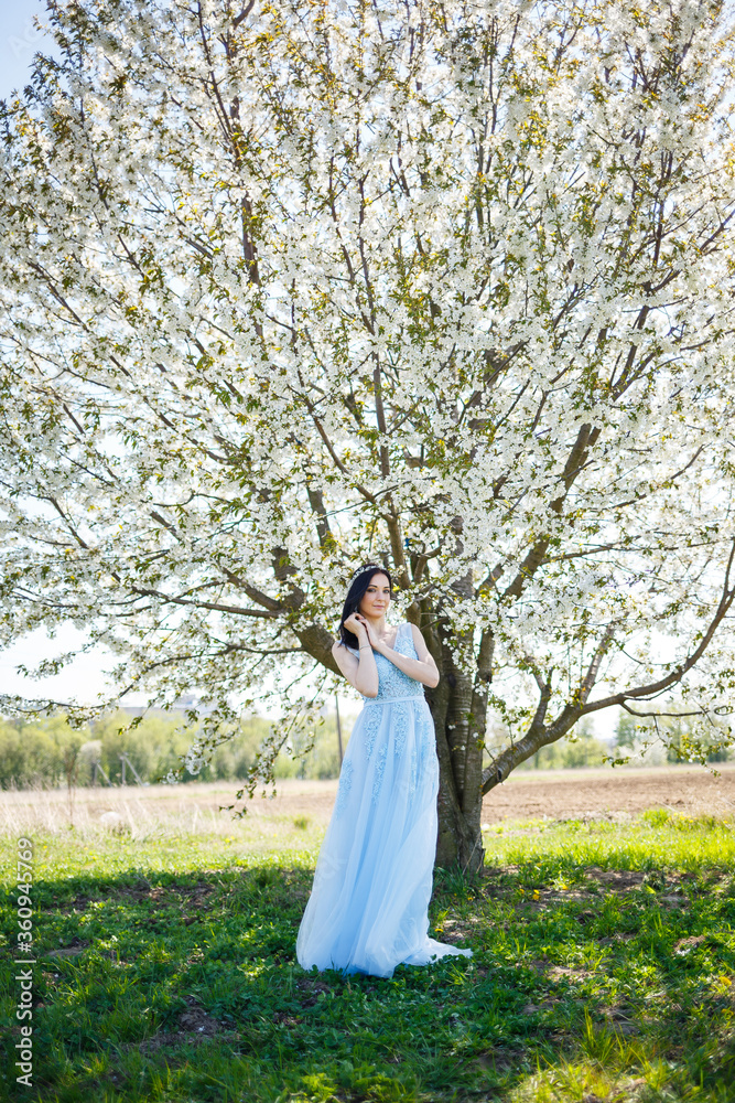 Young woman with beautiful make-up with a vintage hoop in a blue light dress with a bouquet posing on a background of green foliage in a park. Fresh portrait attractive girl with white flowers.