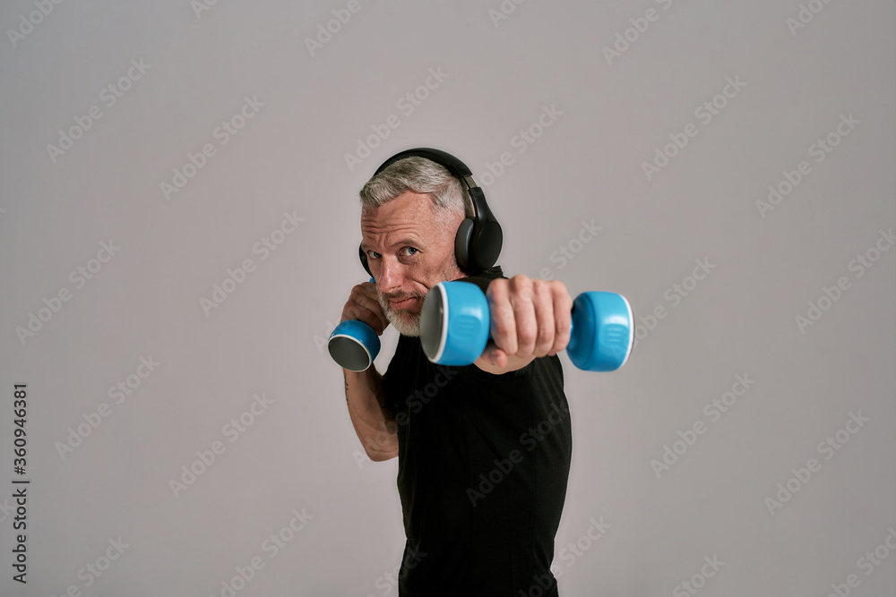 Middle aged muscular man in black t shirt and headphones looking at camera holding blue dumbbells, posing in studio over grey background Middle aged muscular man in black t shirt and headphones looking at camera holding blue dumbbells, posing in studio over grey background