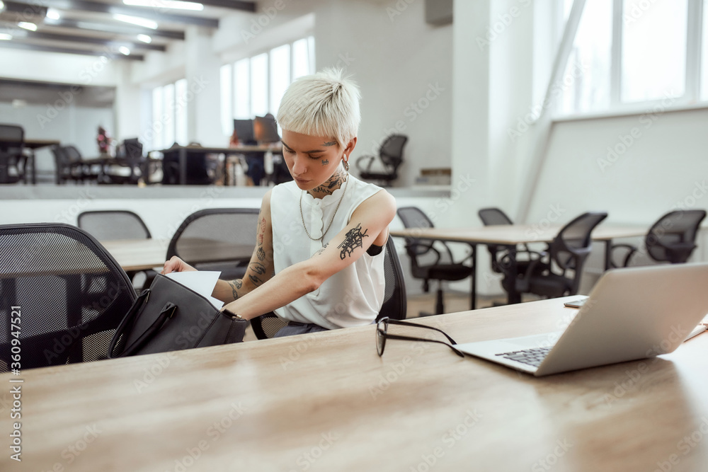 Can not find it. Young cute blonde tattooed businesswoman with short haircut looking for something in her handbag while sitting at her desk in office