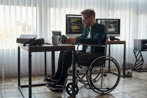 Time to read. Side view of concentrated male web developer in a wheelchair reading a book while sitting at his workplace with multiple computer screens on the background