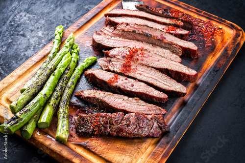 Traditional barbecue sliced dry aged wagyu flank steak offered with green asparagus and chili powder as close-up on a rustic burnt wooden cutting board photo
