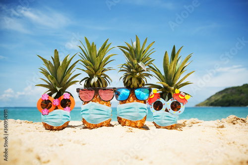 Family of attractive pineapples wearing face mask because of Air pollution or virus epidemic in the world. On the sand against turquoise sea. Summer vacation concept during an epidemic. New reality