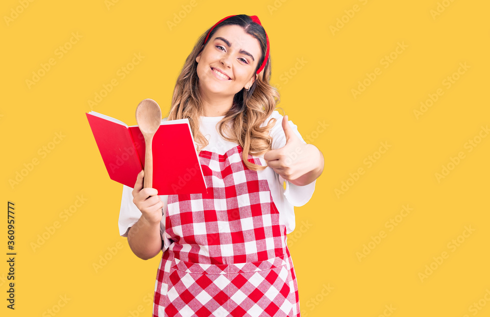Young caucasian woman wearing apron holding recipe book and spoon smiling happy and positive, thumb up doing excellent and approval sign Young caucasian woman wearing apron holding recipe book and spoon smiling happy and positive, thumb up doing excellent and approval sign