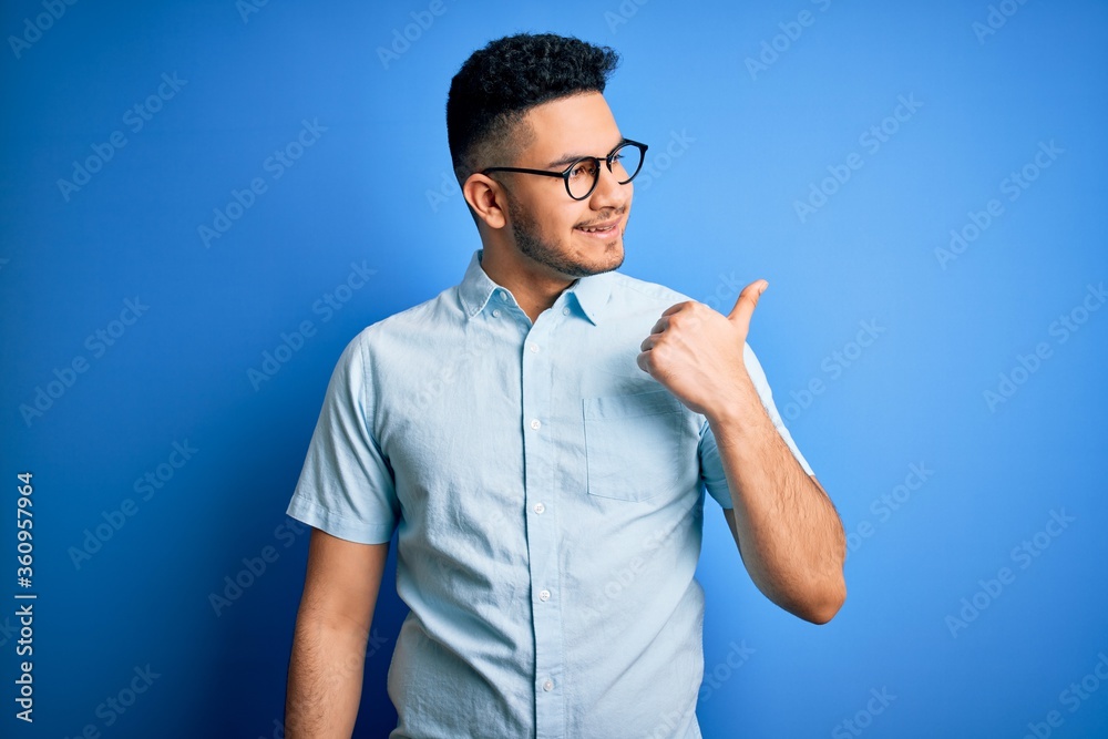 Young handsome man wearing casual summer shirt and glasses over isolated blue background smiling with happy face looking and pointing to the side with thumb up. Young handsome man wearing casual summer shirt and glasses over isolated blue background smiling with happy face looking and pointing to the side with thumb up.