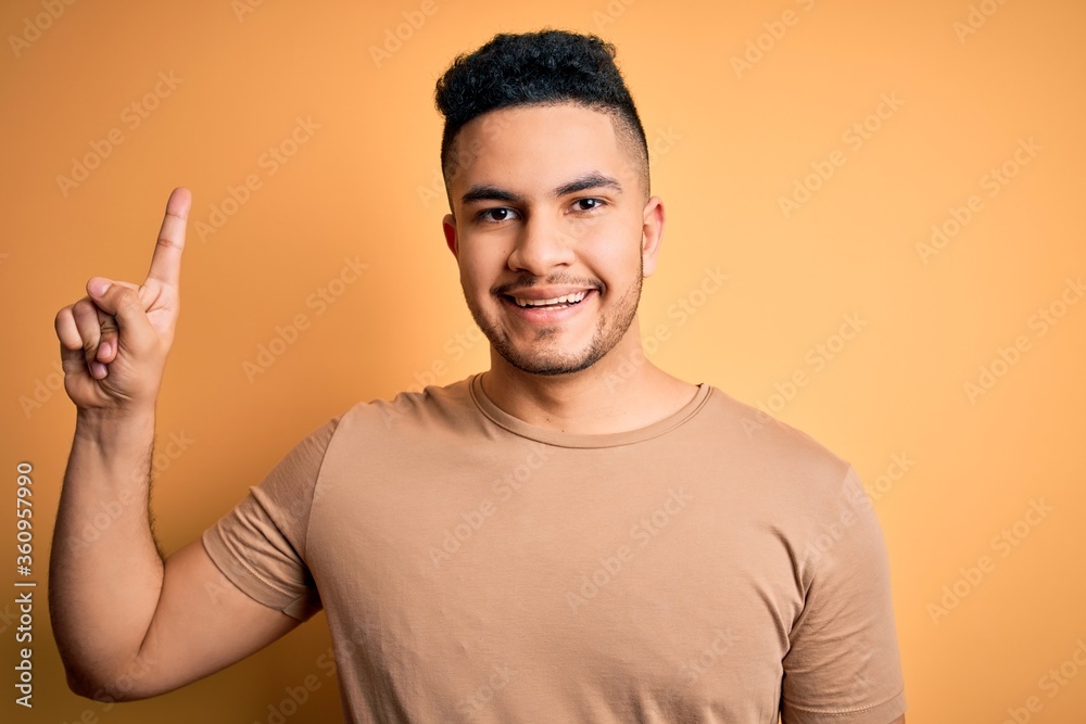 Young handsome man wearing casual t-shirt standing over isolated yellow background showing and pointing up with finger number one while smiling confident and happy. Young handsome man wearing casual t-shirt standing over isolated yellow background showing and pointing up with finger number one while smiling confident and happy.