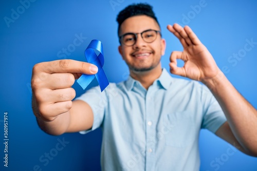 Young handsome man holding blue cancer ribbon standing over isolated background doing ok sign with fingers, excellent symbol