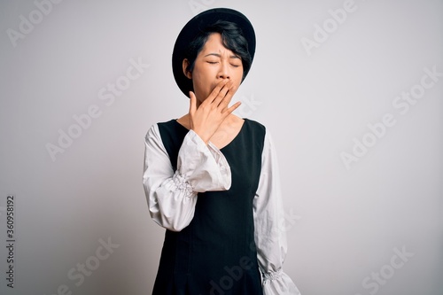 Young beautiful asian girl wearing casual dress and hat standing over isolated white background bored yawning tired covering mouth with hand. Restless and sleepiness.