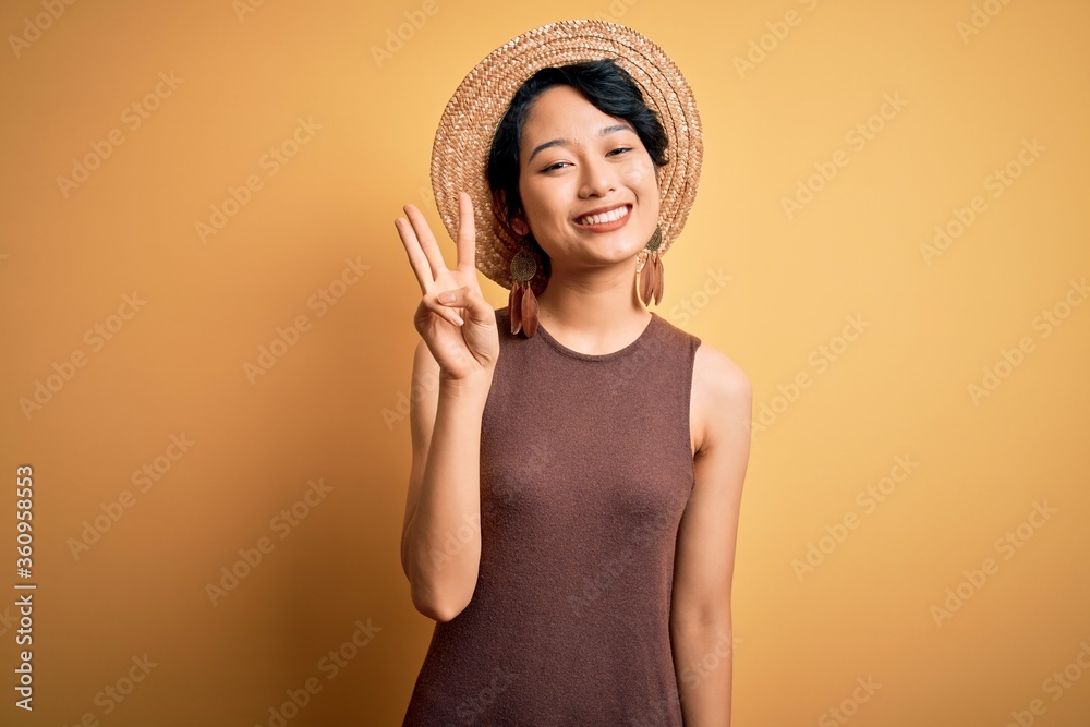 Young beautiful asian girl wearing casual t-shirt and hat over isolated yellow background showing and pointing up with fingers number three while smiling confident and happy. Young beautiful asian girl wearing casual t-shirt and hat over isolated yellow background showing and pointing up with fingers number three while smiling confident and happy.