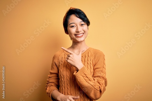 Young beautiful asian girl wearing casual sweater and diadem standing over yellow background cheerful with a smile on face pointing with hand and finger up to the side with happy and natural