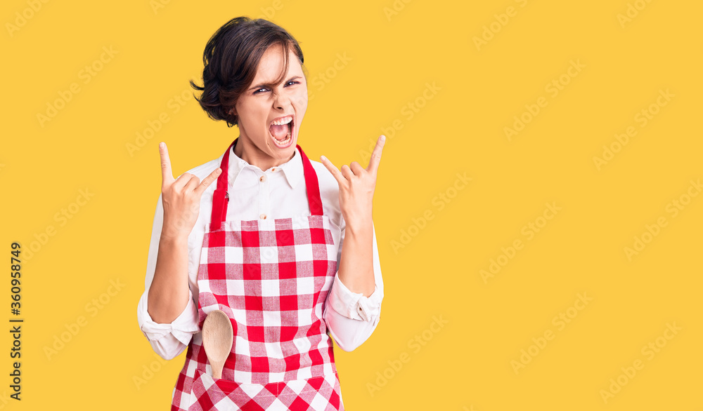 Beautiful young woman with short hair wearing professional cook apron shouting with crazy expression doing rock symbol with hands up. music star. heavy concept. Beautiful young woman with short hair wearing professional cook apron shouting with crazy expression doing rock symbol with hands up. music star. heavy concept.