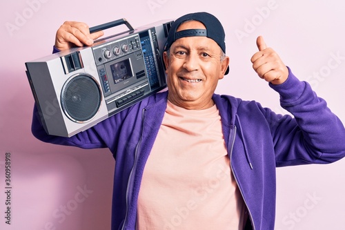 Senior handsome grey-haired modern man listening to music using vintage boombox smiling happy and positive, thumb up doing excellent and approval sign