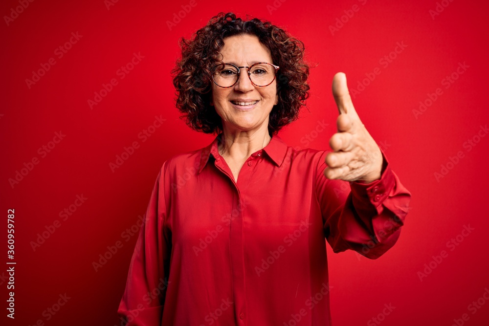 Middle age beautiful curly hair woman wearing casual shirt and glasses over red background smiling friendly offering handshake as greeting and welcoming. Successful business. Middle age beautiful curly hair woman wearing casual shirt and glasses over red background smiling friendly offering handshake as greeting and welcoming. Successful business.