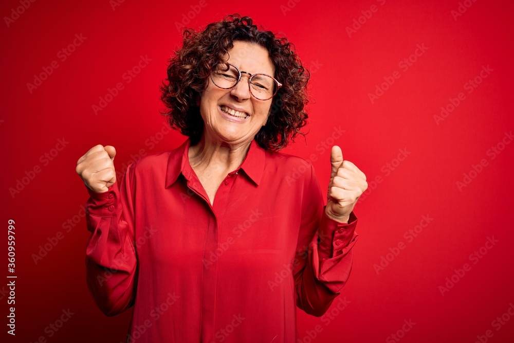 Middle age beautiful curly hair woman wearing casual shirt and glasses over red background very happy and excited doing winner gesture with arms raised, smiling and screaming for success. Celebration