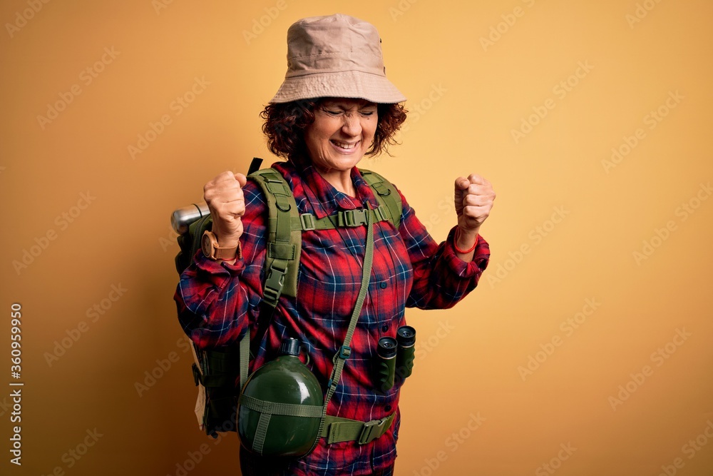 Middle age curly hair hiker woman hiking wearing backpack and water canteen using binoculars very happy and excited doing winner gesture with arms raised, smiling and screaming for success. Celebrate