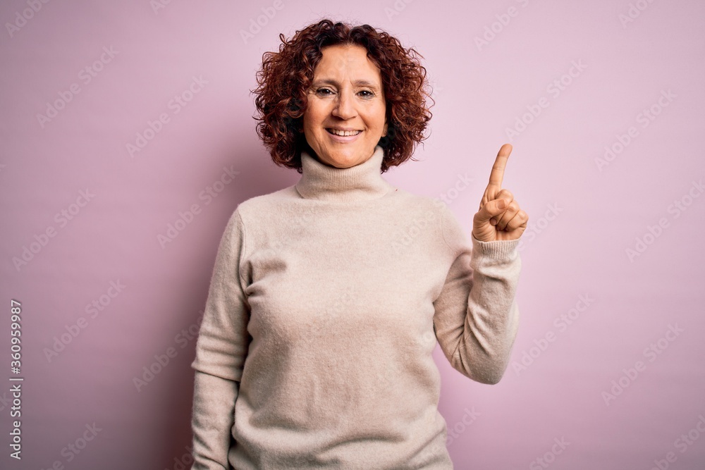 Middle age beautiful curly hair woman wearing casual turtleneck sweater over pink background with a big smile on face, pointing with hand finger to the side looking at the camera. Middle age beautiful curly hair woman wearing casual turtleneck sweater over pink background with a big smile on face, pointing with hand finger to the side looking at the camera.