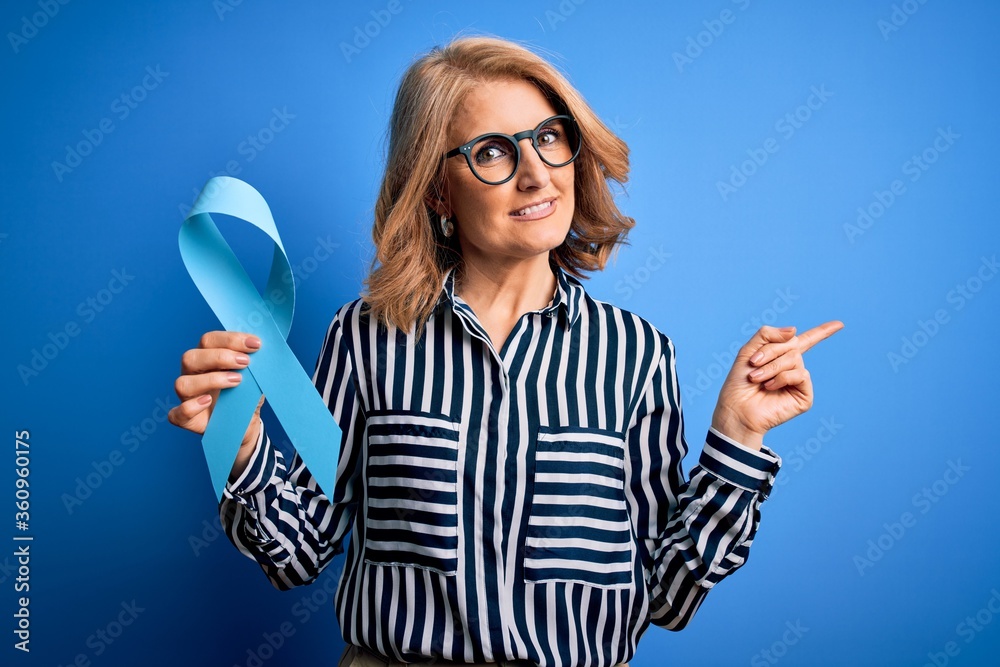 Middle age beautiful blonde woman wearing glasses holding blue cancer ribbon very happy pointing with hand and finger to the side Middle age beautiful blonde woman wearing glasses holding blue cancer ribbon very happy pointing with hand and finger to the side