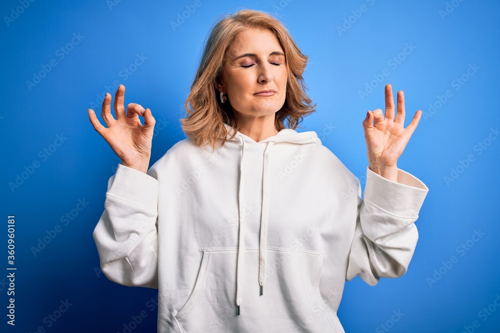 Middle age beautiful blonde sportswoman wearing sportswear over isolated blue background relax and smiling with eyes closed doing meditation gesture with fingers. Yoga concept. Middle age beautiful blonde sportswoman wearing sportswear over isolated blue background relax and smiling with eyes closed doing meditation gesture with fingers. Yoga concept.
