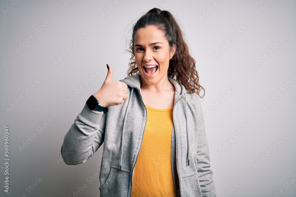 Young beautiful sportswoman with curly hair wearing sportswear over white background doing happy thumbs up gesture with hand. Approving expression looking at the camera showing success. Young beautiful sportswoman with curly hair wearing sportswear over white background doing happy thumbs up gesture with hand. Approving expression looking at the camera showing success.