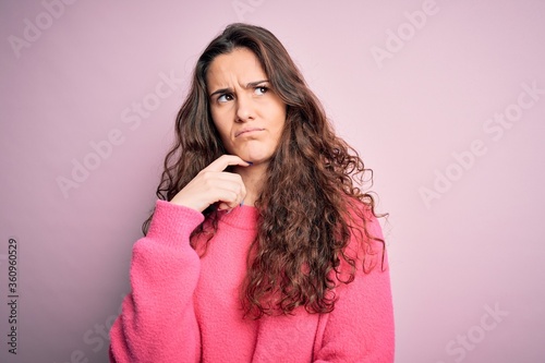Young beautiful woman with curly hair wearing casual sweater over isolated pink background with hand on chin thinking about question, pensive expression. Smiling with thoughtful face. Doubt concept.