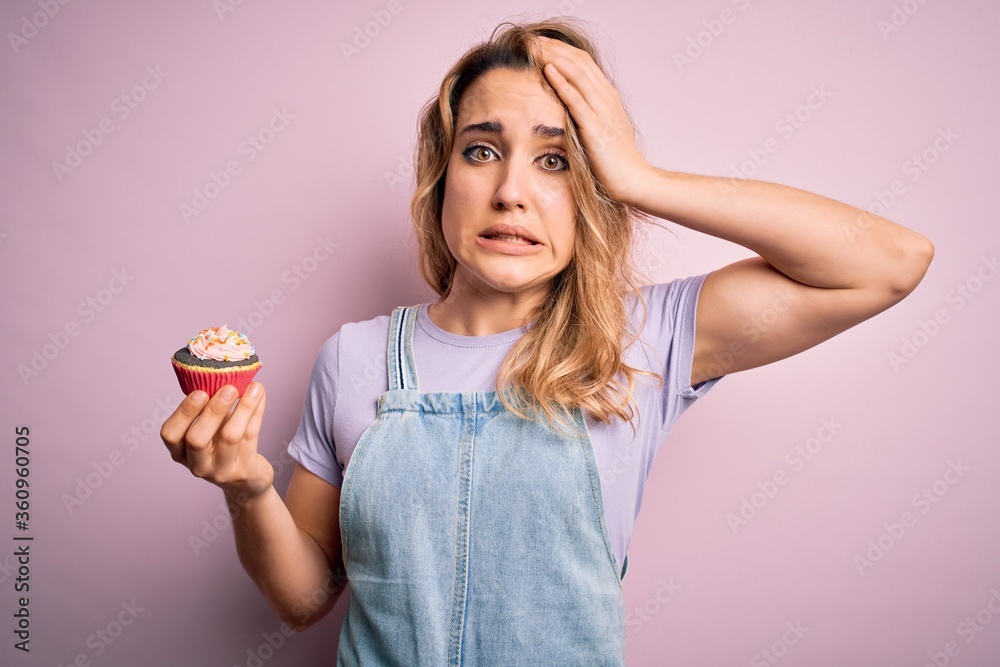 Young beautiful blonde woman eatimg chocolate cupcake over isolated pink background stressed with hand on head, shocked with shame and surprise face, angry and frustrated. Fear and upset for mistake. Young beautiful blonde woman eatimg chocolate cupcake over isolated pink background stressed with hand on head, shocked with shame and surprise face, angry and frustrated. Fear and upset for mistake.