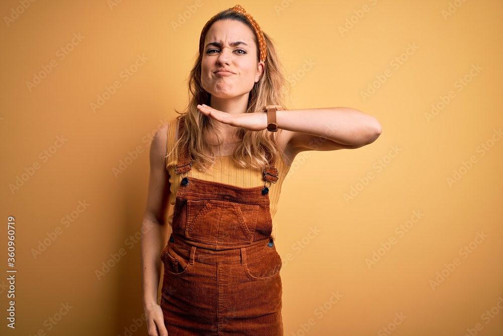 Young beautiful blonde woman wearing overalls and diadem standing over yellow background cutting throat with hand as knife, threaten aggression with furious violence Young beautiful blonde woman wearing overalls and diadem standing over yellow background cutting throat with hand as knife, threaten aggression with furious violence