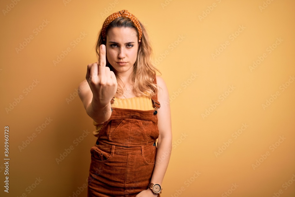 Young beautiful blonde woman wearing overalls and diadem standing over yellow background Showing middle finger, impolite and rude fuck off expression Young beautiful blonde woman wearing overalls and diadem standing over yellow background Showing middle finger, impolite and rude fuck off expression